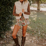 A stylish woman poses outdoors in a cozy fall outfit featuring a white sherpa vest, a brown turtleneck, floral shorts, and knee-high brown boots.