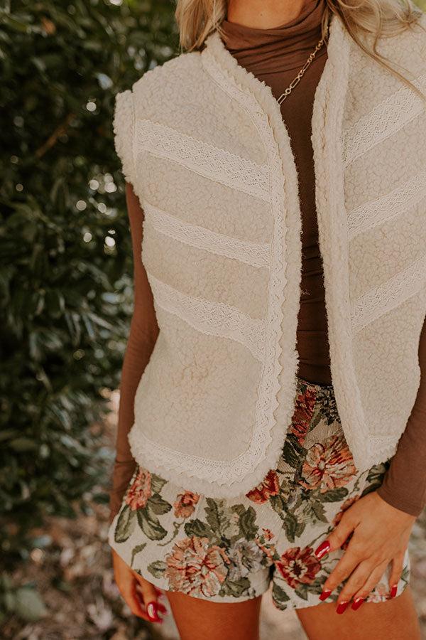 A model dressed in a warm and trendy fall outfit poses on a garden path. Her look features a white sherpa vest, a chocolate brown turtleneck, and floral shorts, perfect for a crisp winter day.