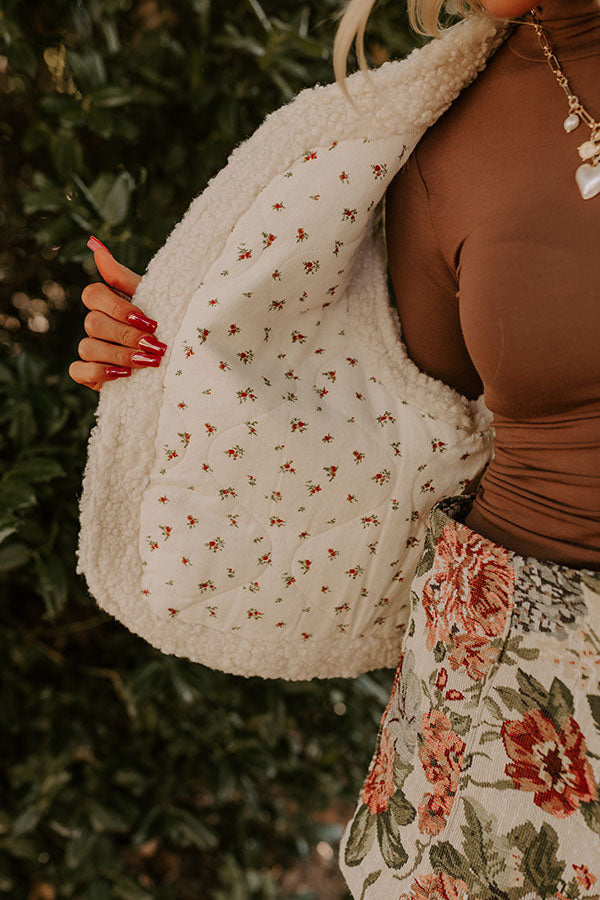 An autumn-inspired outfit includes a cozy white vest over a brown top, and floral-printed shorts. The outdoor background of greenery and sunlight completes the seasonal vibe.