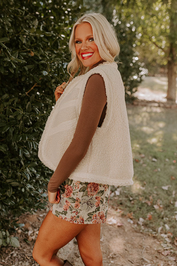 A casual yet elegant fall outfit is styled with a fluffy sherpa vest, a brown turtleneck, and floral-print shorts. The woman accessorizes with brown knee-high boots, posing in a picturesque garden setting.